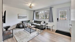 En moderne stue med en grå sofa, et sofabord, et fjernsyn og et stort vindue med gardiner.