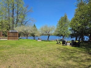 A grassy area with a wooden deck, a small shed, and several Adirondack chairs by a lake. - Stillwater Escape + Hot Tub (Renfrew)
