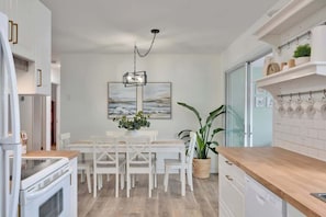 Une cuisine moderne avec des meubles blancs, une table à manger avec des chaises, un luminaire suspendu et une plante en pot.