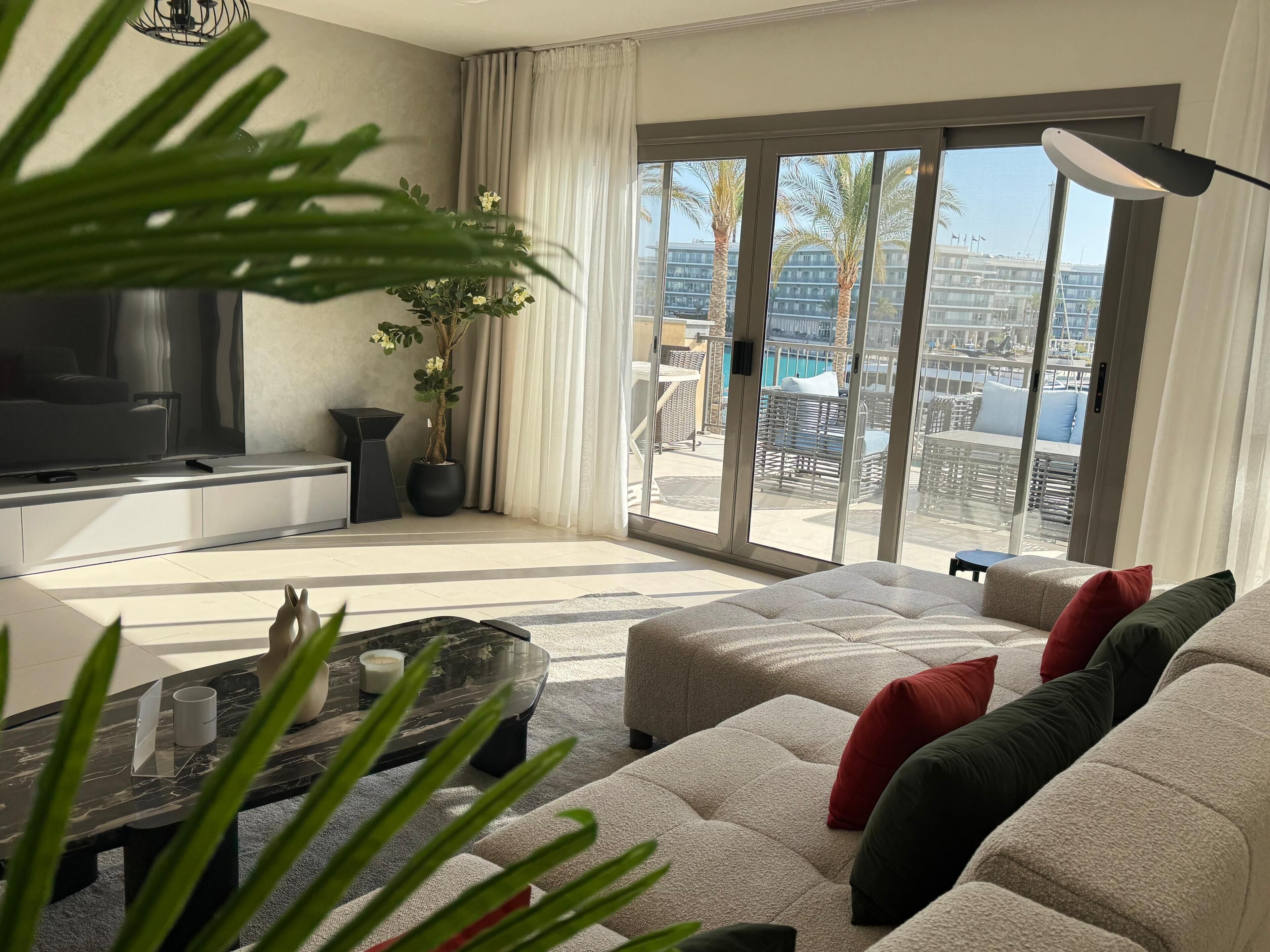 A modern living room with a sofa set, a TV, a potted plant, and a view of a pool and buildings through large glass doors.