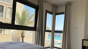 Habitación de hotel con un ventanal grande, una cama, una mesita de noche y vistas a una piscina y edificios.