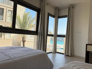 A hotel room with a large window, a bed, a bedside table, and a view of a pool and buildings.