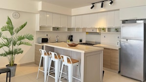 Una cocina moderna con armarios blancos, un refrigerador de acero inoxidable, un microondas y un lavamanos. Hay tres taburetes blancos y una planta en maceta.