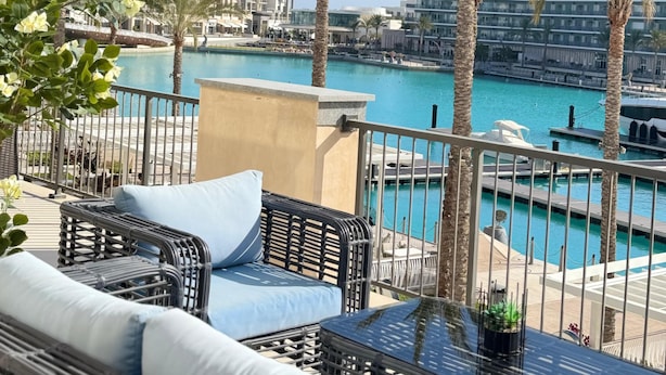 Un balcón con muebles de mimbre, una piscina, palmeras y edificios modernos al fondo.