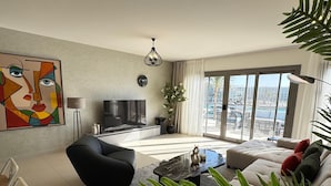 Una sala moderna con un televisor de pantalla plana, un sofá negro, un sillón negro, una mesa de centro y un amplio ventanal con vistas a la ciudad.