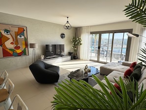 A modern living room with a flat-screen TV, a black sofa, a black armchair, a coffee table, and a large window with a view of a cityscape.