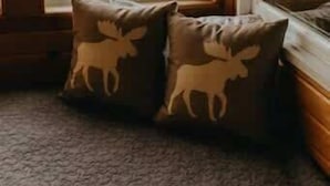 Een kamer met houten wanden, een raam met uitzicht op bomen en een wateroppervlak, en twee kussens met moose afbeeldingen.