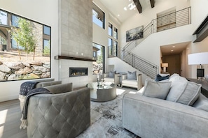 A spacious living room with a fireplace, a staircase, and a large window overlooking a rocky landscape. - 10248 Modane Place-Golf Access & Club Amenities! (Truckee)