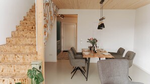 Un interior modern amb una escala de fusta, una zona de menjador amb taula i cadires, i una cuina al fons.