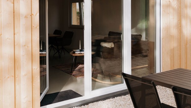 Pared exterior de madera con puerta corredera de vidrio que da a un espacio interior con una mesa y sillas para comer.