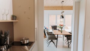 Una cocina moderna con estufa, ollas y cafetera. Hay un comedor con mesa y sillas, y vistas al exterior a través de una ventana.