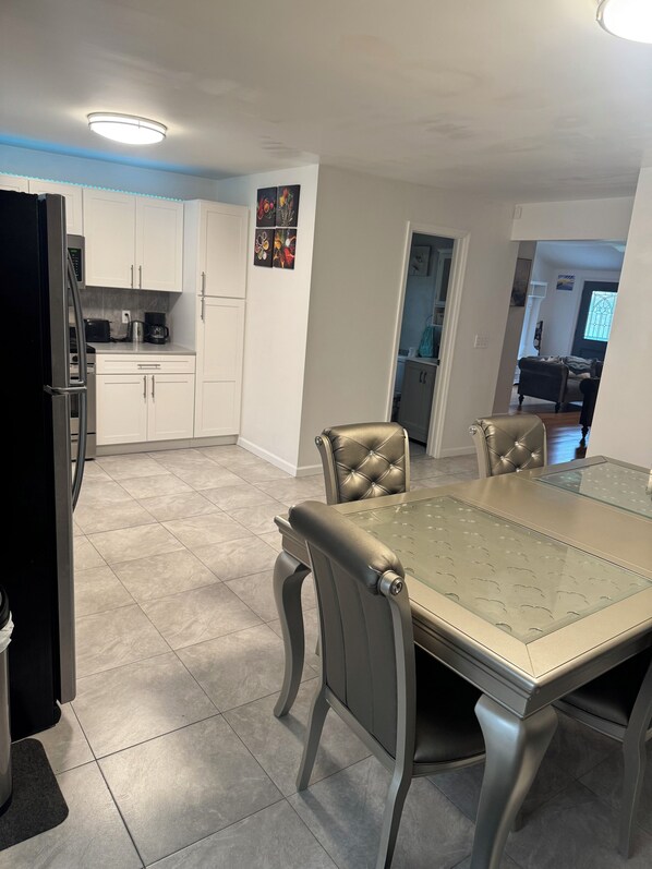 A modern kitchen with white cabinets, a stainless steel refrigerator, and a dining area with a glass-top table and leather chairs.