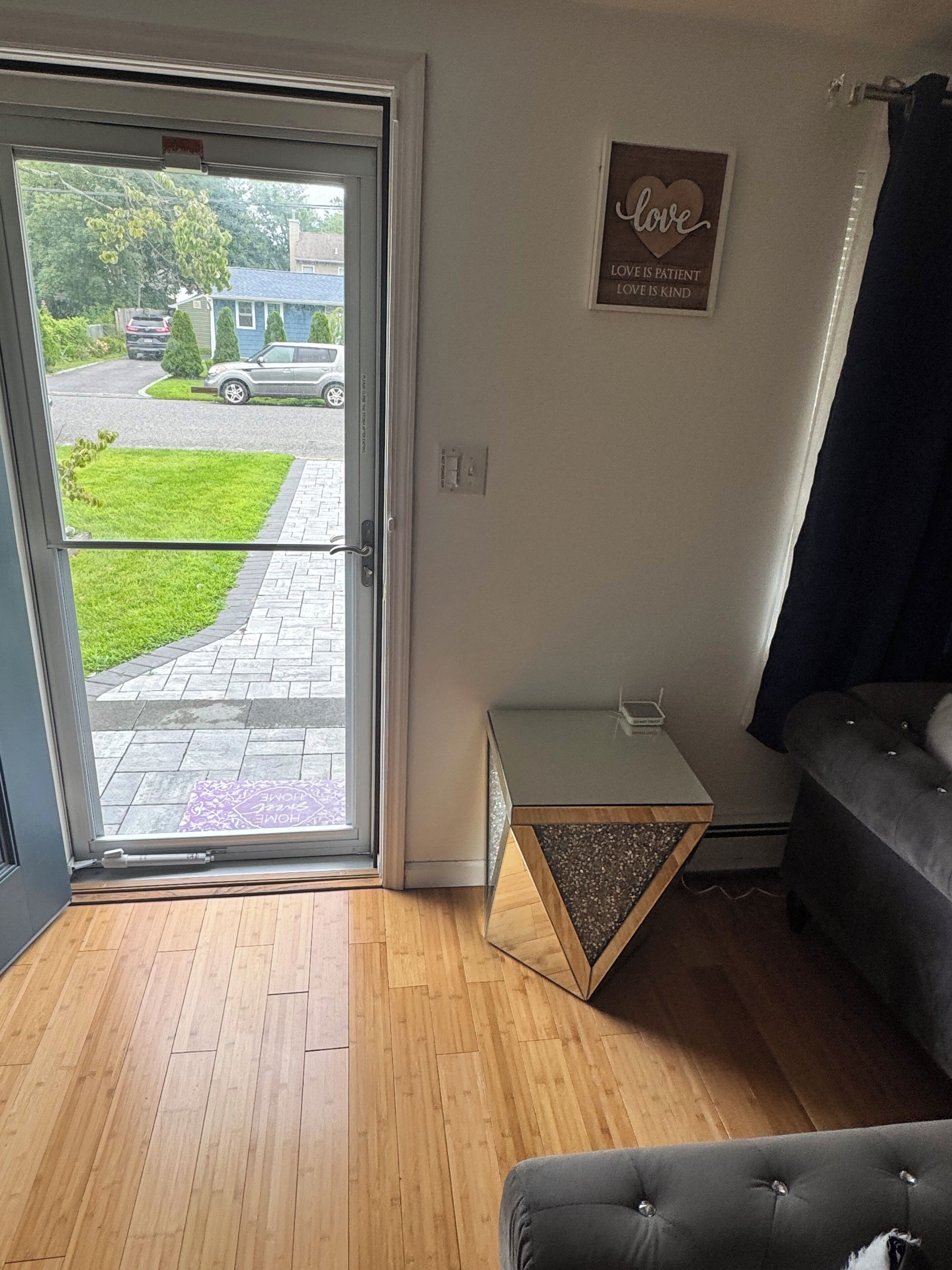 Une pièce dotée d’une porte-fenêtre coulissante donnant sur une allée, d’une petite table d’appoint avec un plateau décoratif, d’un canapé de couleur foncée et d’un panneau mural représentant un cœur et un texte.