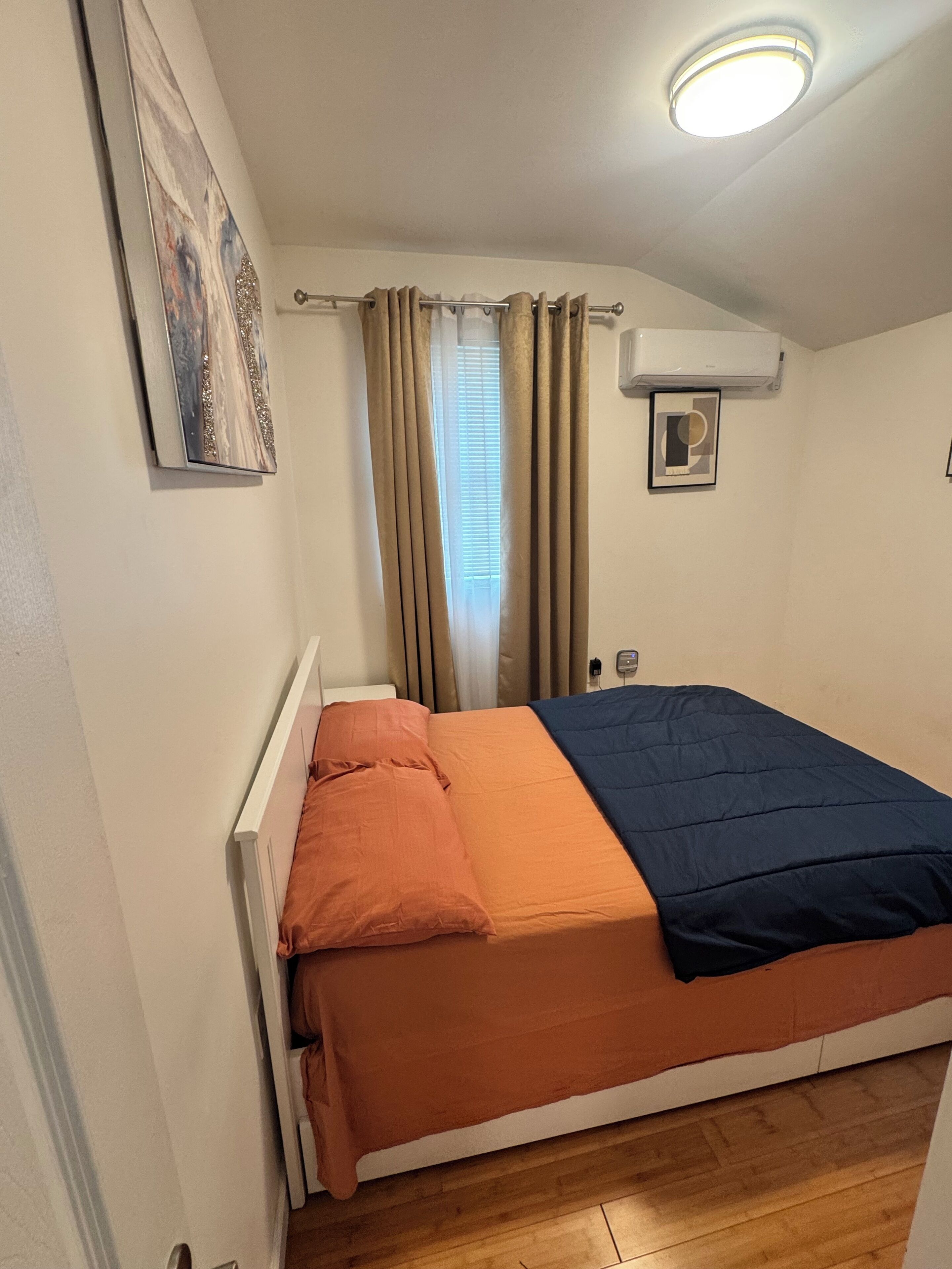 A small, single-bed room with a white headboard, a beige curtain, a ceiling light, and a wall-mounted air conditioning unit.