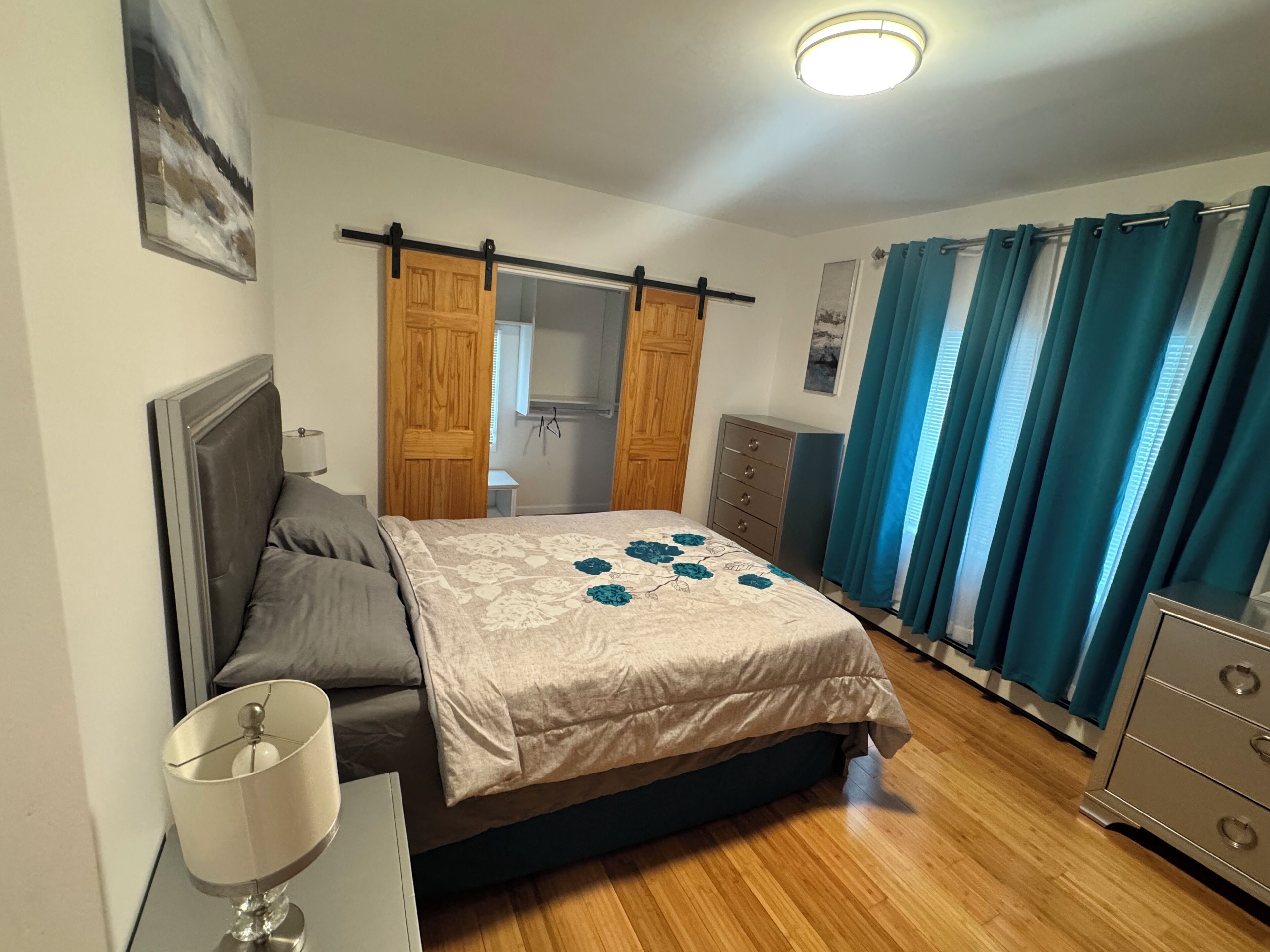 A bedroom with a bed, a dresser, a nightstand, a lamp, a wardrobe, and a ceiling light.