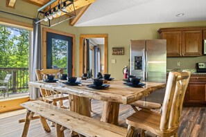 Ein Holzes Esstisch mit Stühlen, ein Küchenbereich mit Schränken, einem Kühlschrank und Blick ins Freie.