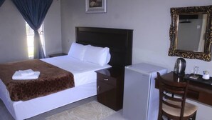 A hotel room with a bed, a desk, a refrigerator, a chair, a mirror, and a window with curtains. - The Empire Inn (Serowe)