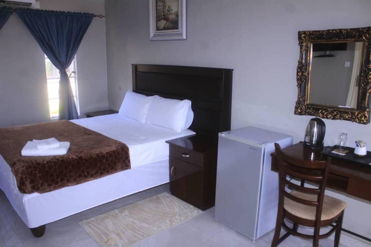 A hotel room with a bed, a desk, a refrigerator, a chair, a mirror, and a window with curtains.