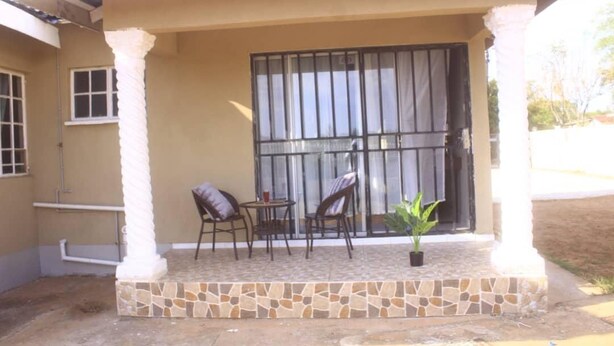 A covered outdoor seating area with stone flooring, two chairs, a small table, and a potted plant. - The Empire Inn (Serowe)