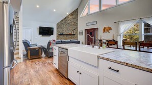 A modern kitchen with a large island, a dining area with wooden chairs, a stone accent wall, and a TV mounted on the wall. - Cranmore Heights | Luxe, Hot Tub, Walk Downtown (North Conway)
