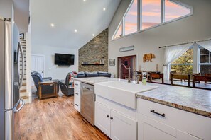 A modern kitchen with a large island, a dining area with wooden chairs, a stone accent wall, and a TV mounted on the wall.