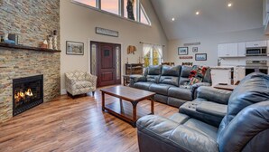 A spacious living room with a stone fireplace, leather sofas, a wooden coffee table, and a kitchen area with white cabinets. - Cranmore Heights | Luxe, Hot Tub, Walk Downtown (North Conway)