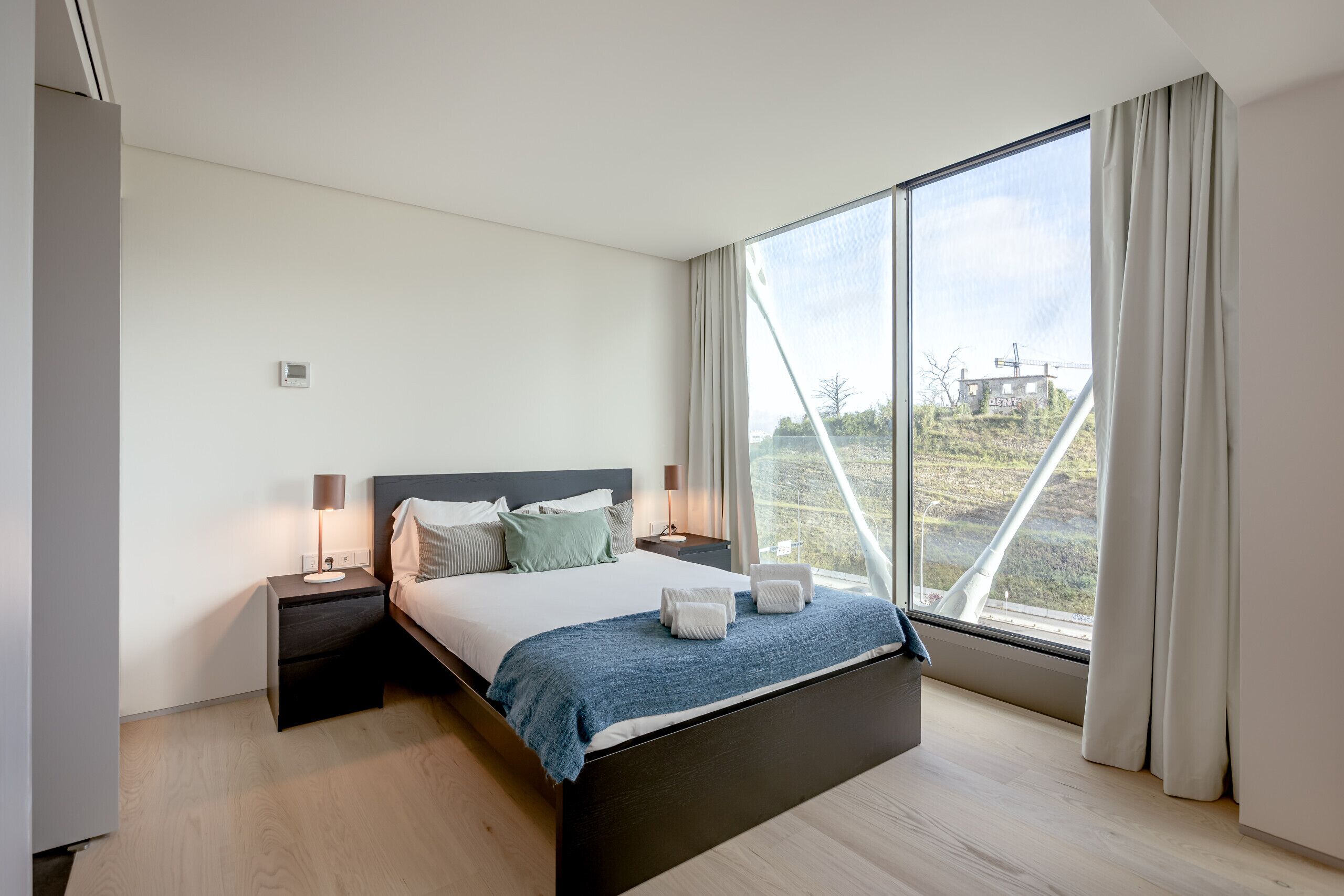 A modern bedroom with a large bed, bedside tables, a view through a large window, and a view of the outside landscape.