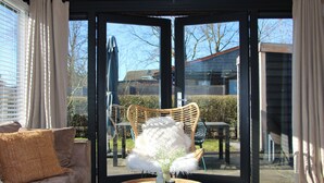 A room with a sofa, a chair, a table, and a view through a glass door. - The Kingfisher is a comfortable ground-floor bungalow with fenced garden, wood stove and fully equipped kitchen. Near the beach, dunes and forest of Schoorl, Petten and Bergen. Perfect for families and dog lovers. (Dirkshorn)