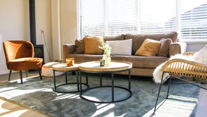 A living room with a sofa, two coffee tables, a chair, and a large window with blinds. - The Kingfisher is a comfortable ground-floor bungalow with fenced garden, wood stove and fully equipped kitchen. Near the beach, dunes and forest of Schoorl, Petten and Bergen. Perfect for families and dog lovers. (Dirkshorn)