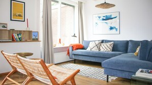Una sala de estar con un sofá azul, un sillón tejido, una estantería y un amplio ventanal con cortinas.