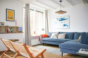 Ein Wohnzimmer mit einem blauen Sofa, einem gewobenen Sessel, einem Bücherregal und einem großen Fenster mit Vorhängen.