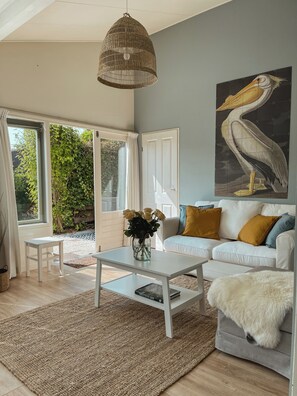 A living room with a white sofa, a coffee table, a hanging light fixture, and a large pelican painting on the wall. - The Pelican is a recently renovated ground-floor holiday bungalow on the ’t Geestmerambacht park near Schoorl and Bergen. Enjoy peace, sunshine and nature close to the beach, dunes, forest and tulip fields. (Warmenhuizen)