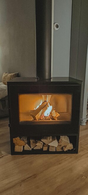 A wood-burning stove with a glass front, a black chimney, and logs stored below. - The Pelican is a recently renovated ground-floor holiday bungalow on the ’t Geestmerambacht park near Schoorl and Bergen. Enjoy peace, sunshine and nature close to the beach, dunes, forest and tulip fields. (Warmenhuizen)