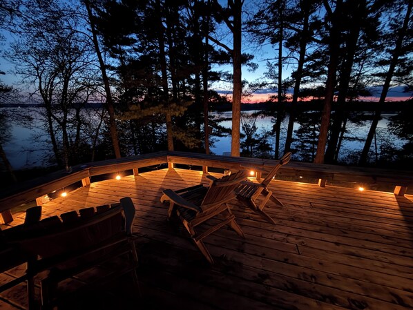 A wooden deck with chairs and a fire pit, surrounded by trees and overlooking a body of water.