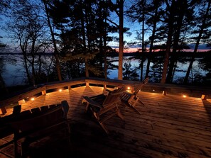Una terraza de madera con sillas y una chimenea, rodeada de árboles y con vista a un cuerpo de agua.