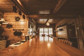 Een houten interieur met een lange eettafel, een keukenhoek en grote ramen.