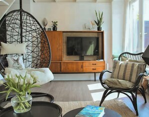 A living room with a TV, wooden cabinet, wicker chair, and a book titled 'Van Gogh'. - A boutique coastal lodge between sea and forest, personally managed by Dutch Sweet Home. With a fireplace, sunny garden and serene atmosphere — created for slow living and true relaxation. (Warmenhuizen)