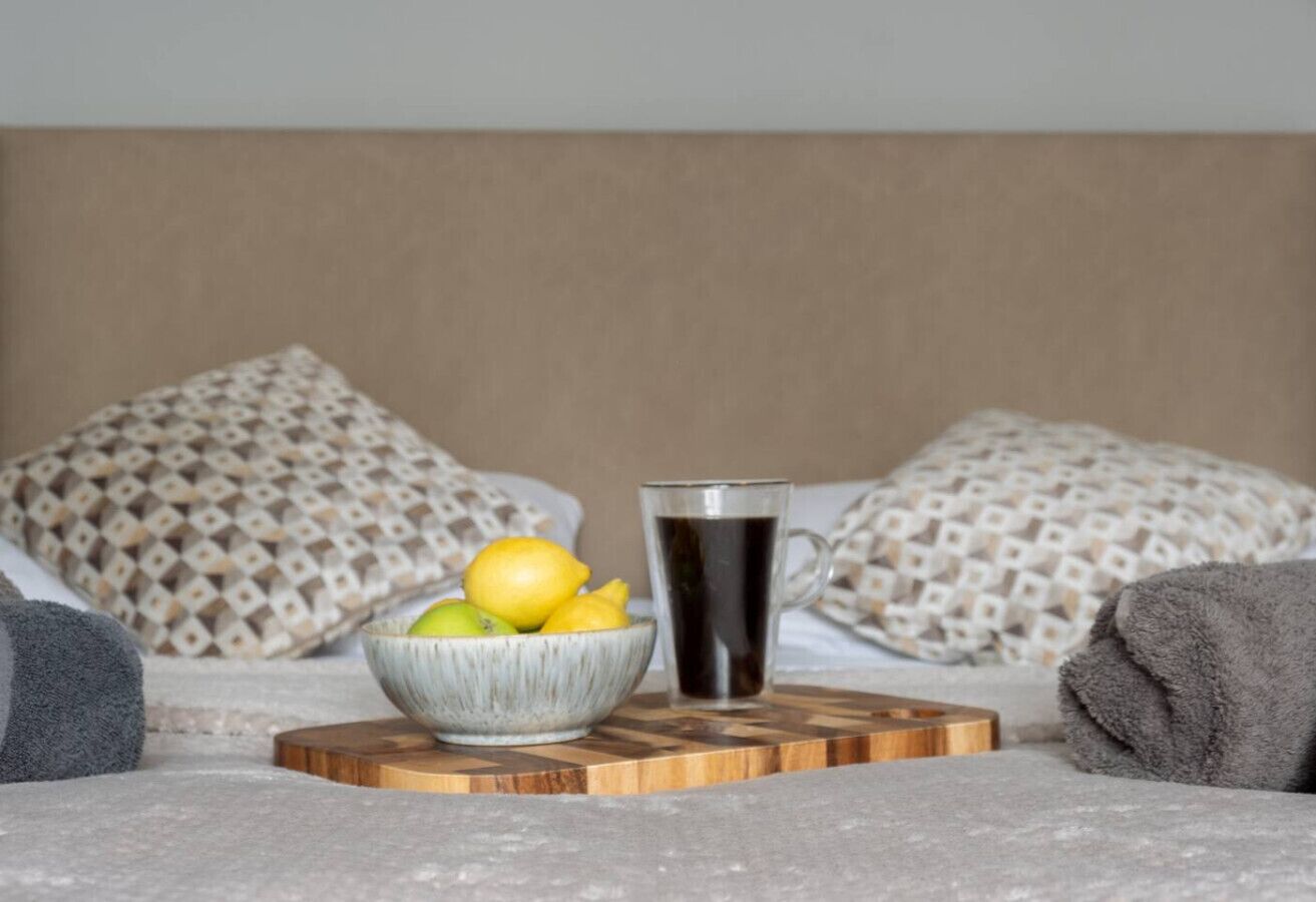 A bed with patterned pillows, a bowl of fruit, and a coffee mug on a wooden coaster.