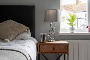 Une chambre à coucher avec un lit, une table de chevet avec une lampe, un verre d’eau et une plante en pot près d’une fenêtre.