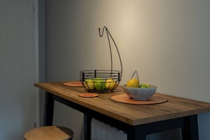 Une table de salle à manger en bois, sur laquelle se trouvent deux bols remplis de fruits, un panier métallique contenant des fruits et des dessous de verre.
