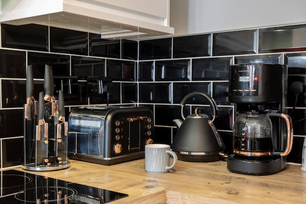 Une cuisine avec un plan de travail recouvert de carreaux noirs, une machine à café, une bouilloire, un grille-pain et une tasse posée sur le plan de travail.