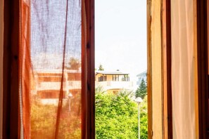 Blick durch ein Fenster mit Vorhängen, der einen Gebäudebalkon und umliegendes Grün zeigt.
