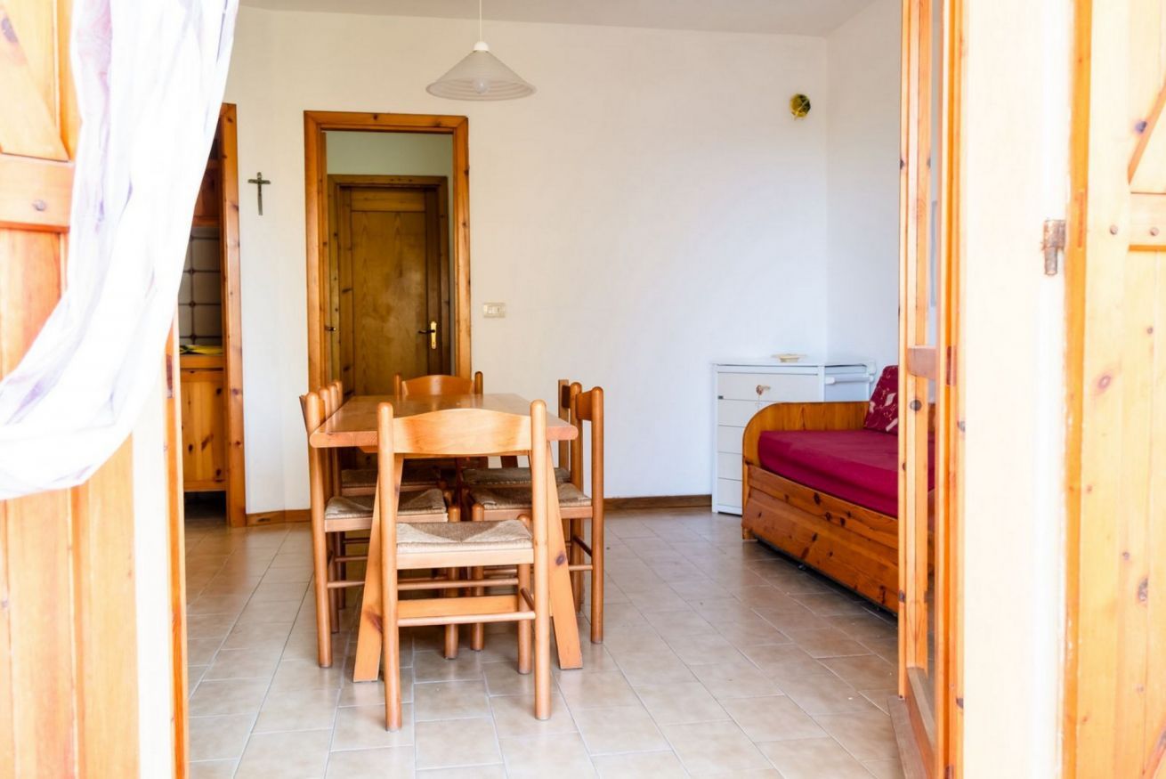 A room with a wooden dining table, chairs, a bed with a red blanket, and a small white cabinet.