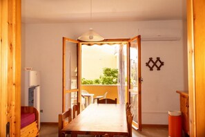 Une pièce comprenant une table à manger en bois, des chaises, une fenêtre avec des rideaux, un climatiseur blanc et une petite table avec une chaise à l’extérieur.