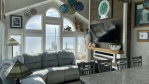 A modern living room with a large sofa, a dining area with chairs, a TV, and a view of the sea through large windows. - Sandbridge Beach, Coastal Chic Back Bay Waterfront Tiny Home! (Virginia Beach)