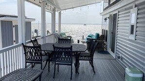 A covered deck with outdoor furniture, a view of the water, and a building with windows. - Sandbridge Beach, Coastal Chic Back Bay Waterfront Tiny Home! (Virginia Beach)