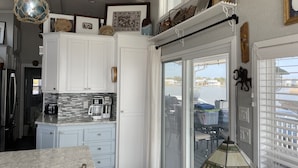 A kitchen with white cabinets, a coffee maker, and a view of a body of water through a sliding glass door. - Sandbridge Beach, Coastal Chic Back Bay Waterfront Tiny Home! (Virginia Beach)