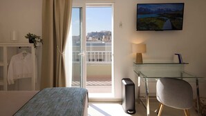 Hotellihuone, jossa on sänky, työpöytä, tuoli, lamppu, taulu ja suuri ikkuna, josta avautuu näkymä kaupunkiin.