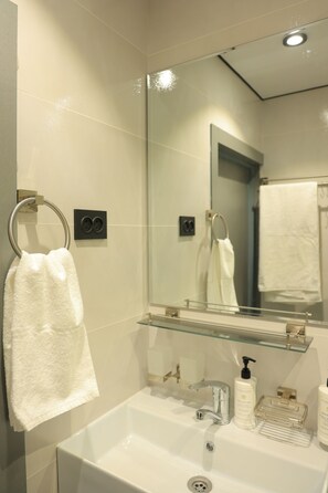 Un baño con lavamanos blanco, un espejo, un perchero para toallas y un estante de vidrio con artículos de aseo.