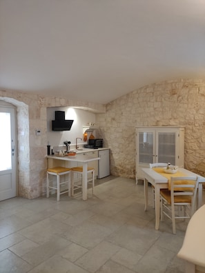 A compact kitchen with a stone wall, a microwave, a sink, and a small refrigerator. There is a dining table with chairs and a cabinet with glass doors. - Complex of trulli and lamie in Itria Valley - charm and relaxation by the pool (Puglia)